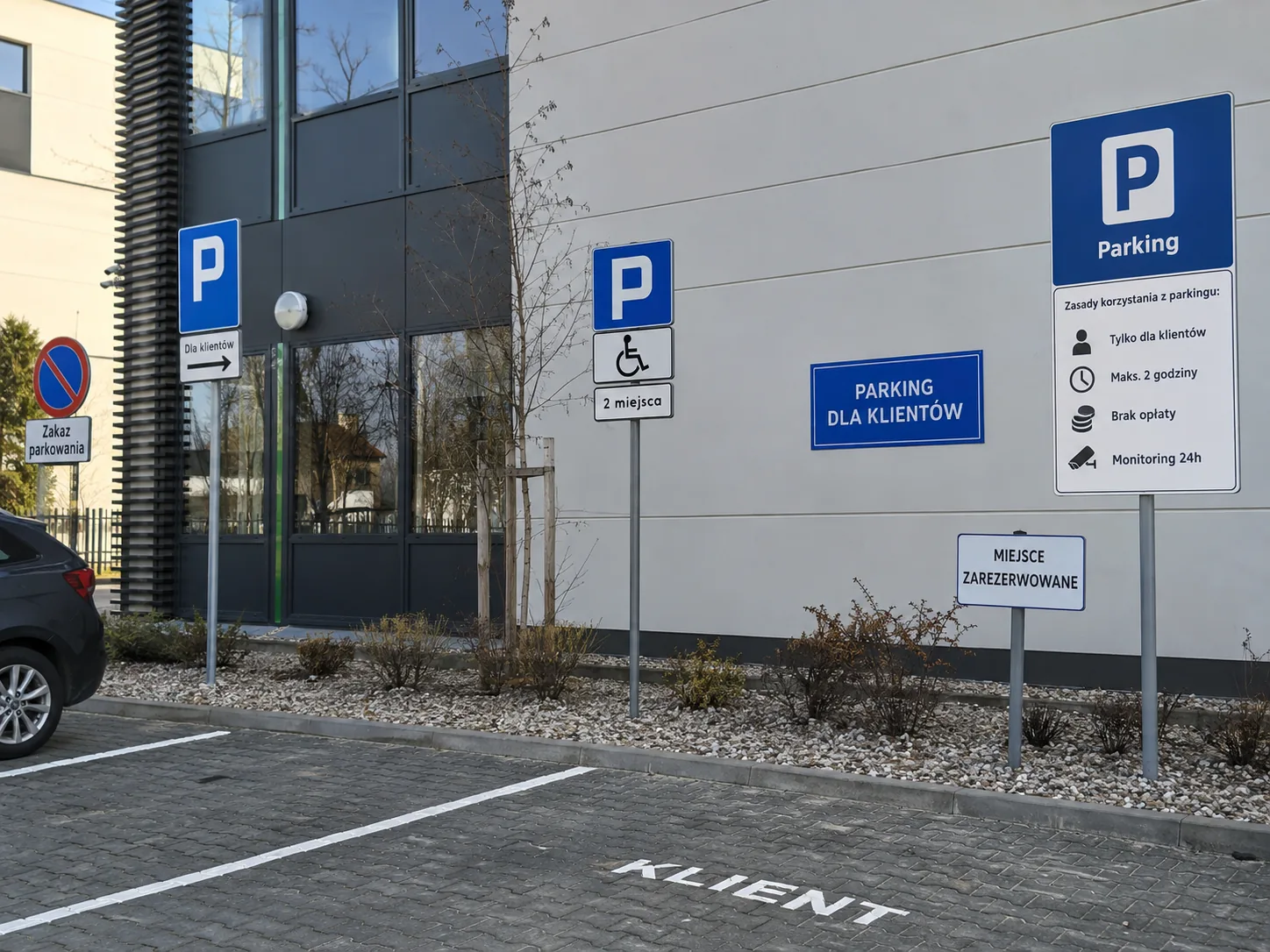 Oznaczenia parkingowe przy budynku firmy: znaki P na słupkach, tablica z regulaminem parkingu i tabliczka miejsca zarezerwowanego